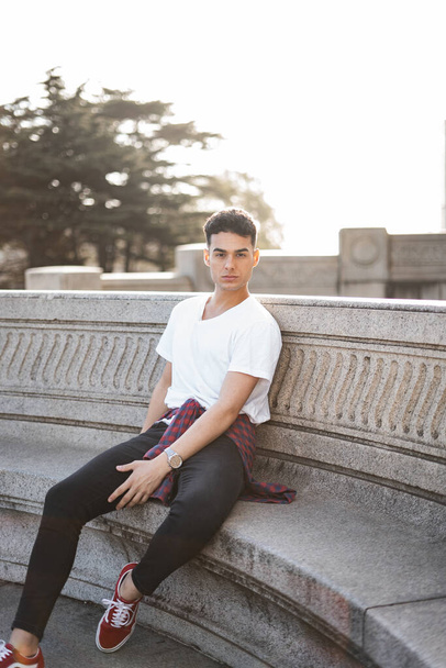 A vertical shot of a young stylish male in a white t-shirt sitting on a stone bench - Fotoğraf, Görsel