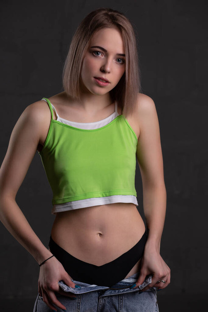 Gorgeous sexy blonde girl in a crop top and unbuttoned jeans poses on a dark background looking at the camera - Zdjęcie, obraz