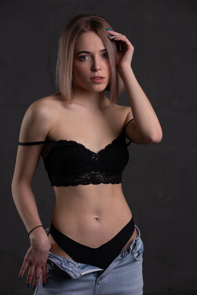 Beautiful young blonde woman in a black bra, panties and stylish jeans. Her gorgeous shoulders are bare. Photo on a dark background - Foto, afbeelding