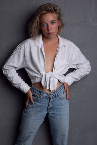 a gorgeous young blonde woman in an unbuttoned shirt and jeans poses on a gray background - Foto, Bild