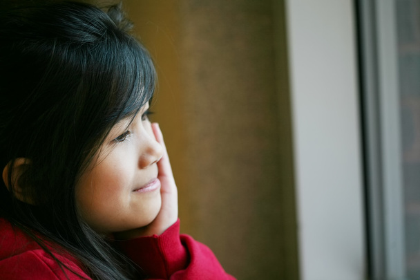 Little Asian girl quietly sitting by window, hand on chin - Photo, Image