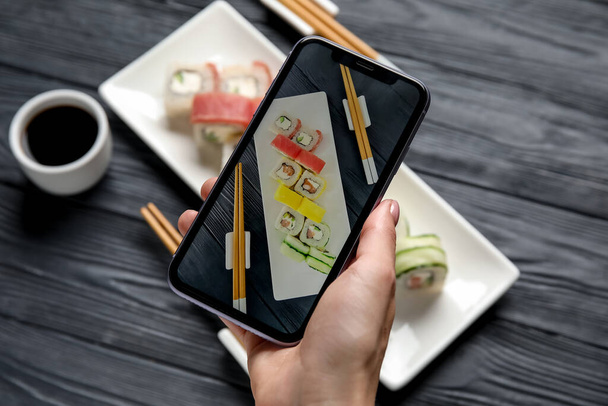 Mujer sosteniendo el teléfono móvil y tomando una foto de la placa con un conjunto de deliciosos rollos de sushi en la mesa de madera gris - Foto, imagen