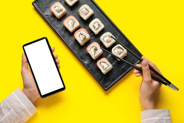 Woman holding mobile phone and eating delicious sushi rolls on color background - Fotó, kép