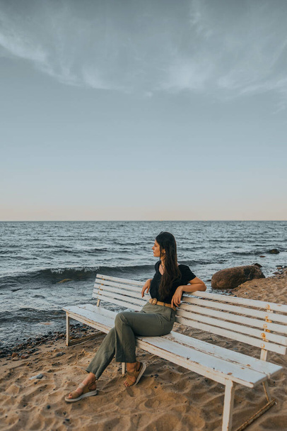 Calm girl with dark hair is sitting on the white bench on the seashore. - Photo, Image