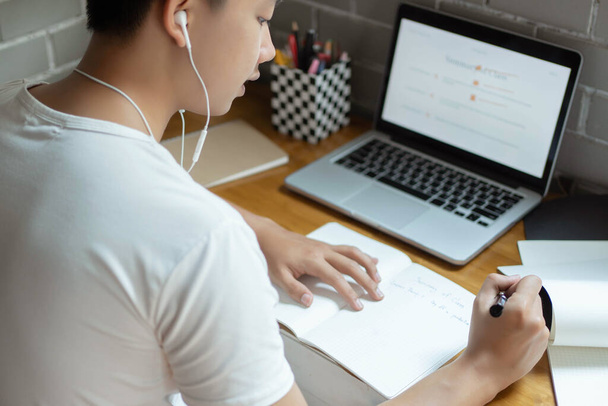 Online σπουδάζοντας concept ο μαθητής λυκείου ακούγοντας μουσική μέσω των ακουστικών του και χρησιμοποιώντας το laptop του για να αναζητήσει πληροφορίες σχετικά με την εργασία του στην εφημερίδα. - Φωτογραφία, εικόνα