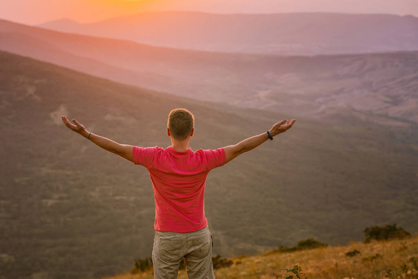 Genç sarışın bir adam gün batımında dağın tepesini fethetti ve elleri havada duruyor. Eğimli güzel bir dağ manzarası. - Fotoğraf, Görsel