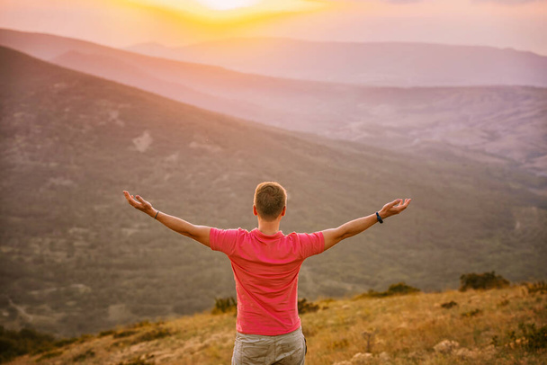 Zadní pohled mladý plavovlasý muž dobyl vrchol hory při západu slunce a stojí se zdviženýma rukama. Krásná horská krajina se svahem. - Fotografie, Obrázek