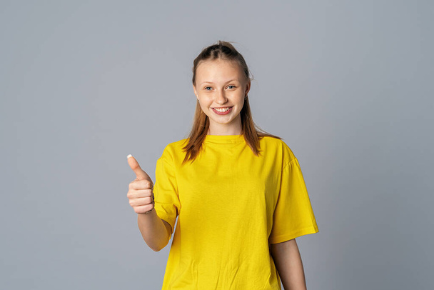 Beautiful smiling teen girl, showing thumb up isolated on light gray background, dressed in casual yellow t shirt - Foto, Imagen