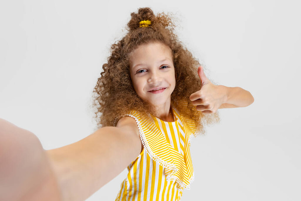 Front camera view of little smiling curly beautiful girl looking at camera isolated over white studio background. - Photo, Image