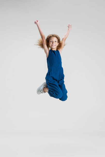 One little Caucasian cute girl in blue stylish jumpsuit jumping high and having fun isolated over white studio background. - Фото, изображение