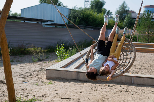 Mom and daughter swing on a round swing. Caucasian woman and little girl have fun on the playground. - Φωτογραφία, εικόνα