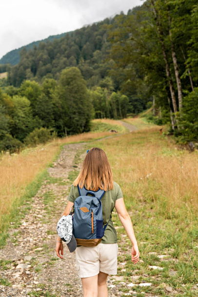 Vista trasera de la joven rubia con mochila caminando cuesta arriba a lo largo del sendero en el fondo de la montaña cubierta de bosque y nubes cielo, estilo de vida saludable activa senderismo ecoturismo - Foto, Imagen