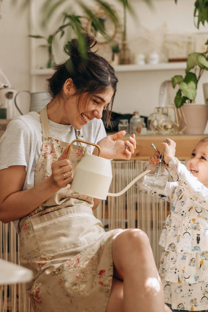 Mixed race family young woman and her little daughter is watering houseplants at home and having fun splashing water.Home gardening.Family leisure,hobby concept.Biophilia design and urban jungle concept - Photo, image