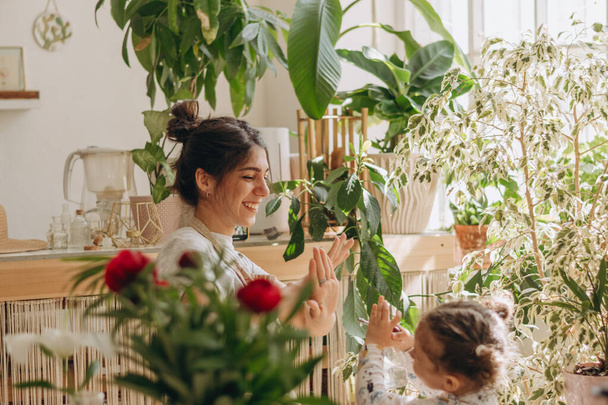Mixed race family woman with her little daughter is watering houseplants at home and having fun splashing water.Home gardening.Family leisure,hobby concept.Biophilia design and urban jungle concept. - Photo, image