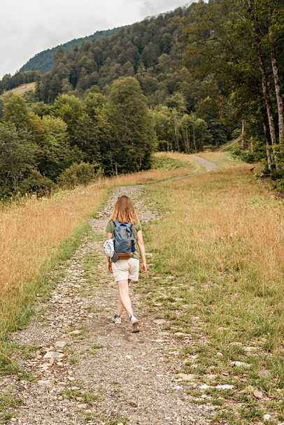 Rear view of young blonde woman with backpack walking uphill along trail on background of mountain covered with forest and clouds sky, active healthy lifestyle hiking eco tourism - Fotó, kép