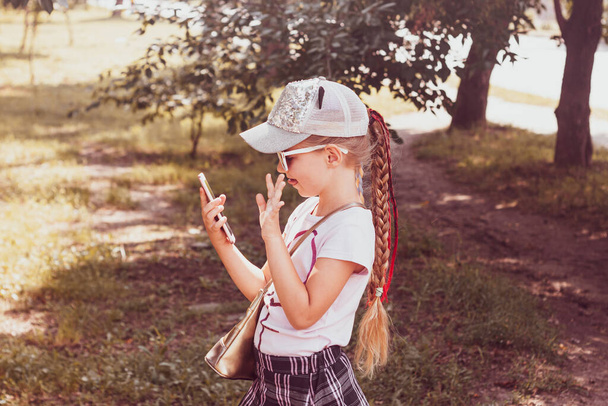 Чарівна маленька дівчинка з довгим плетеним волоссям у бейсбольній шапці зі смартфоном на відкритому повітрі осінь сонячний день. Молодіжна культура спосіб життя. Щасливі дитячі листи в осінньому парку. Автентична шкільна дитина ходить
. - Фото, зображення