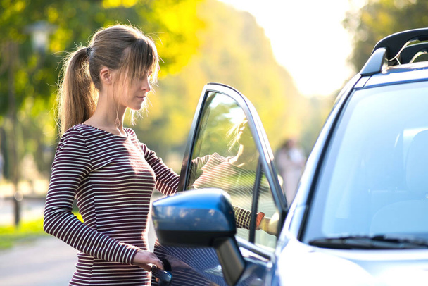 Fiatal boldog női sofőr áll a kocsija mellett egy városi utcában nyáron. Utazási célpontok és szállítási koncepció. - Fotó, kép