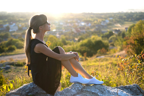 Fiatal elegáns nő fekete rövid ruhában ül egy sziklán pihentető szabadban nyáron este. Divatos nő élvezi a meleg naplementét a természetben. - Fotó, kép