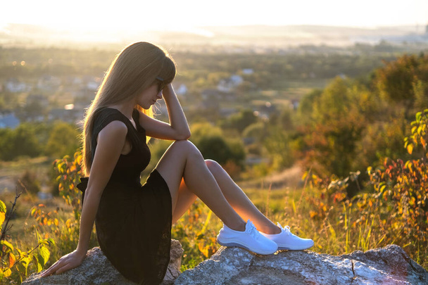 Jolie femme triste en robe d'été courte noire assise sur un rocher pensant à l'extérieur au coucher du soleil. Femme à la mode contemplant dans une soirée chaude dans la nature. - Photo, image
