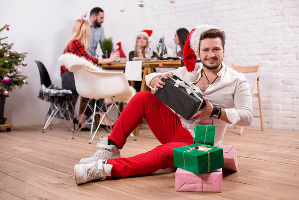 Shot of happy friends enjoying holidays. Focus on the man with a gift boxes in the foreground in a red Christmas hat - Photo, Image