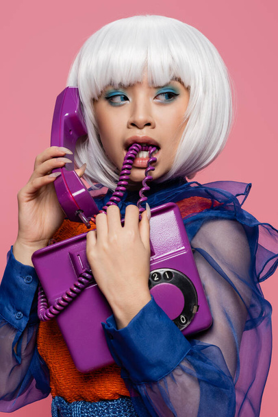 Ασιατική ποπ τέχνη μοντέλο δάγκωμα καλώδιο, ενώ μιλάμε στο τηλέφωνο απομονώνονται σε ροζ  - Φωτογραφία, εικόνα