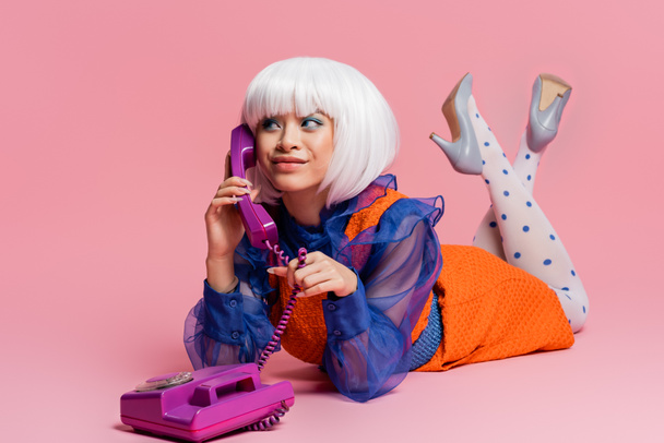 Sonriente asiático pop arte modelo hablando por teléfono mientras que la mentira en rosa fondo - Foto, Imagen