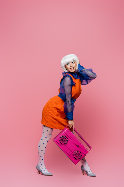 Cheerful asian woman in white wig and retro dress holding boombox on pink background - Fotó, kép