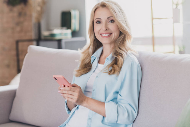Photo of cute pretty age woman dressed blue shirt sitting sofa typing modern device smiling indoors home room - Valokuva, kuva
