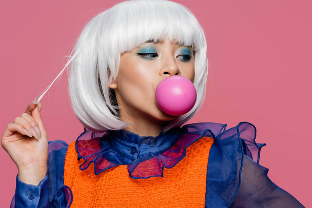 Stylish asian woman touching hair and blowing bubble gum isolated on pink  - Photo, Image