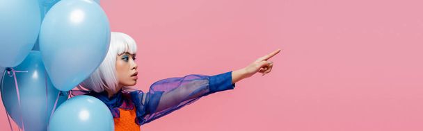 Κομψό ασιατικό μοντέλο pop art δείχνοντας με το δάχτυλο κοντά μπαλόνια που απομονώνονται σε ροζ, banner  - Φωτογραφία, εικόνα