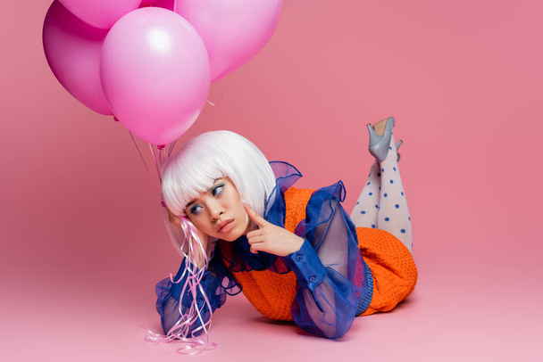 Pensive asian pop art woman holding balloons while lying on pink background - Foto, Imagem