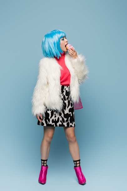 Full length of stylish asian pop art woman eating donut on blue background  - Фото, изображение