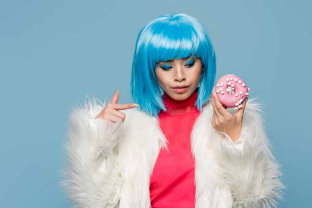Stylish asian woman in fluffy jacket pointing at donut isolated on blue  - Foto, afbeelding