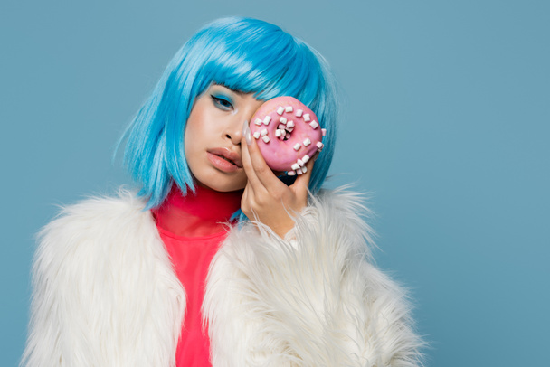 Trendy young asian woman in fluffy jacket holding donut near face isolated on blue  - Foto, afbeelding