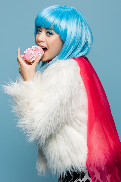 aziatische vrouw in pop art stijl eten donut en kijken naar camera geïsoleerd op blauw  - Foto, afbeelding