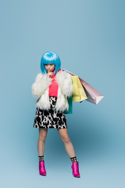 Mujer asiática en estilo pop art sosteniendo bolsas de compras y mostrando gesto secreto sobre fondo azul - Foto, Imagen