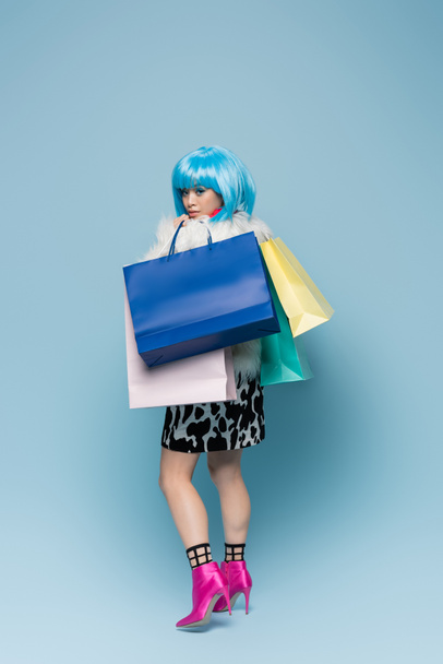 Trendy asian woman in heels and fluffy jacket holding shopping bags on blue background  - Photo, Image
