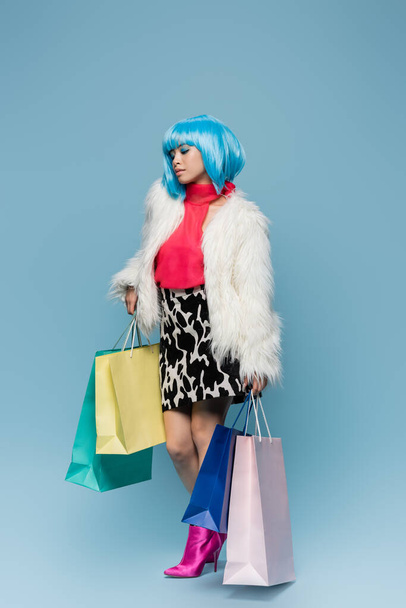 Pretty asian woman in bright wig holding colorful shopping bags on blue background  - Valokuva, kuva