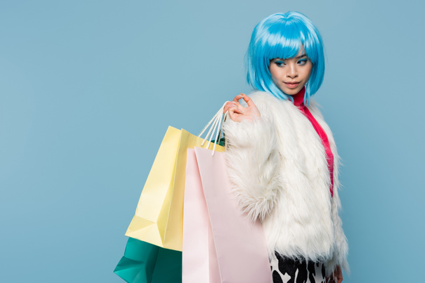 Modelo asiático en estilo pop art posando con bolsas de compras aisladas en azul  - Foto, Imagen