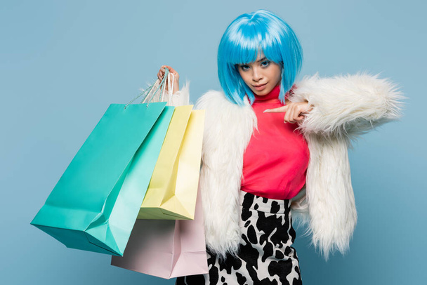 Stylish asian woman in bright wig pointing at shopping bags isolated on blue  - Фото, изображение
