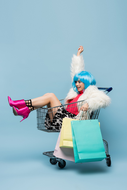 Cheerful asian pop art woman holding shopping bags while sitting in cart on blue background  - Photo, Image