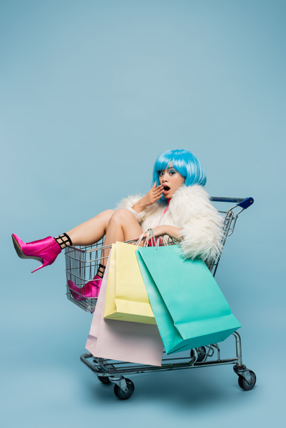 Εκπληκτική Ασιάτισσα γυναίκα σε στυλ pop art κοιτάζοντας κάμερα, ενώ κρατώντας τσάντες για ψώνια στο καλάθι σε μπλε φόντο  - Φωτογραφία, εικόνα
