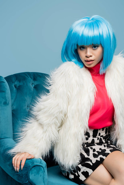 Stylish asian model in wig and fluffy jacket sitting on armchair isolated on blue  - 写真・画像