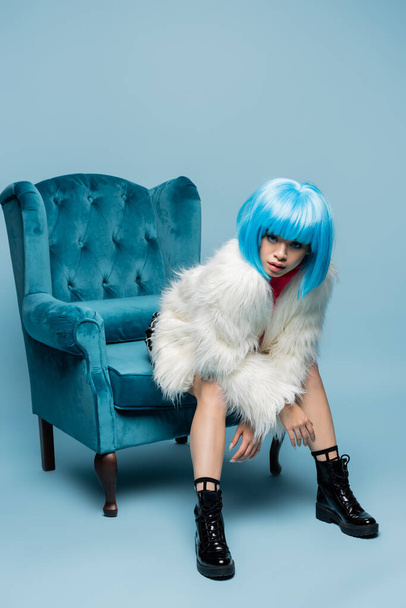 Asian woman in bright wig and fluffy jacket looking at camera on turquoise armchair on blue background  - Foto, Imagem