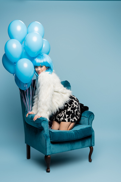 Smiling asian pop art model looking at camera near balloons on armchair on blue background  - Fotografie, Obrázek