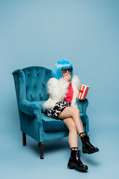 Stylowa azjatycka pop art kobieta w okularach przeciwsłonecznych jedzenie popcorn w fotelu na niebieskim tle  - Zdjęcie, obraz