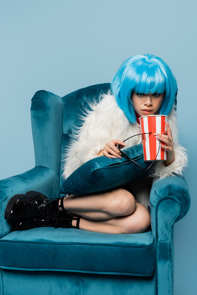 Stylish asian pop art woman holding sunglasses and popcorn on armchair isolated on blue  - Foto, afbeelding
