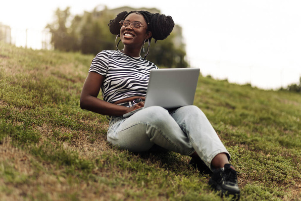 Portrait of attractive cheerful black focused woman , sitting on grass with laptop while spending free time outdoors - Fotografie, Obrázek