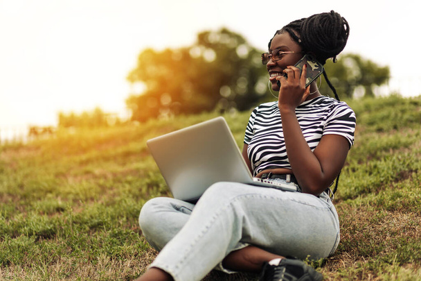 Portrait of smiling black young woman wearing stylish glasses using laptop and talking on mobile phone outdoors, concept of technology, business, communication - Фото, изображение