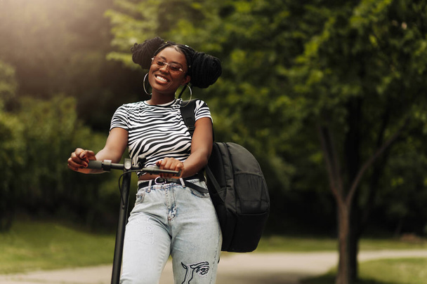 Happy black female student, with a backpack, rides an electric scooter to the university, outdoors in the park, modern woman, new generation, electric transport, ecology, ecological transport - Photo, image